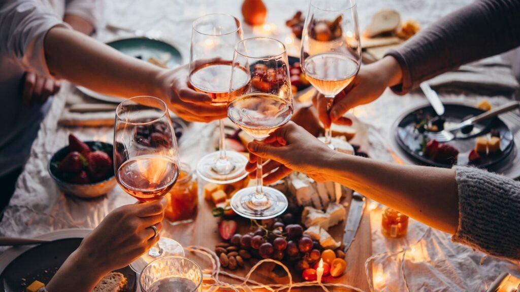 Close-up of friends raising glasses of rosé wine in a toast over a dinner table filled with charcuterie, fruit, candles, and festive lights, creating a cozy and elegant atmosphere.