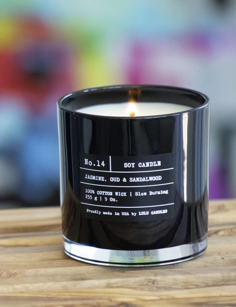Lit soy candle in a sleek black glass container labeled Jasmine, Oud & Sandalwood, sitting on a wooden surface with a colorful blurred background.